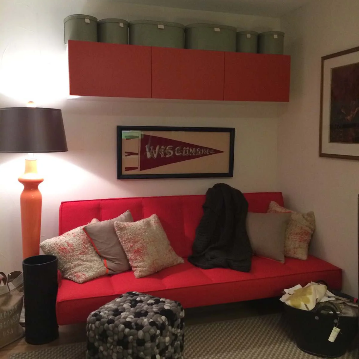 Red sofa bed beneath wall storage with a small felted ottoman and a tall orange lamp