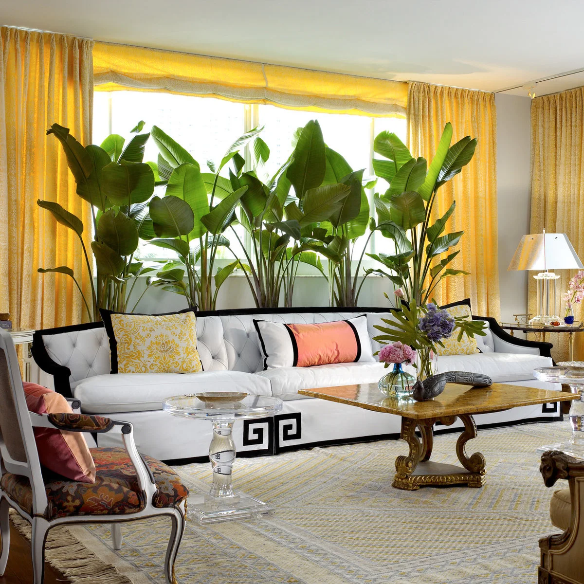 White sofa beneath yellow drapes with black trim, patterned cushions and large leafy plants behind