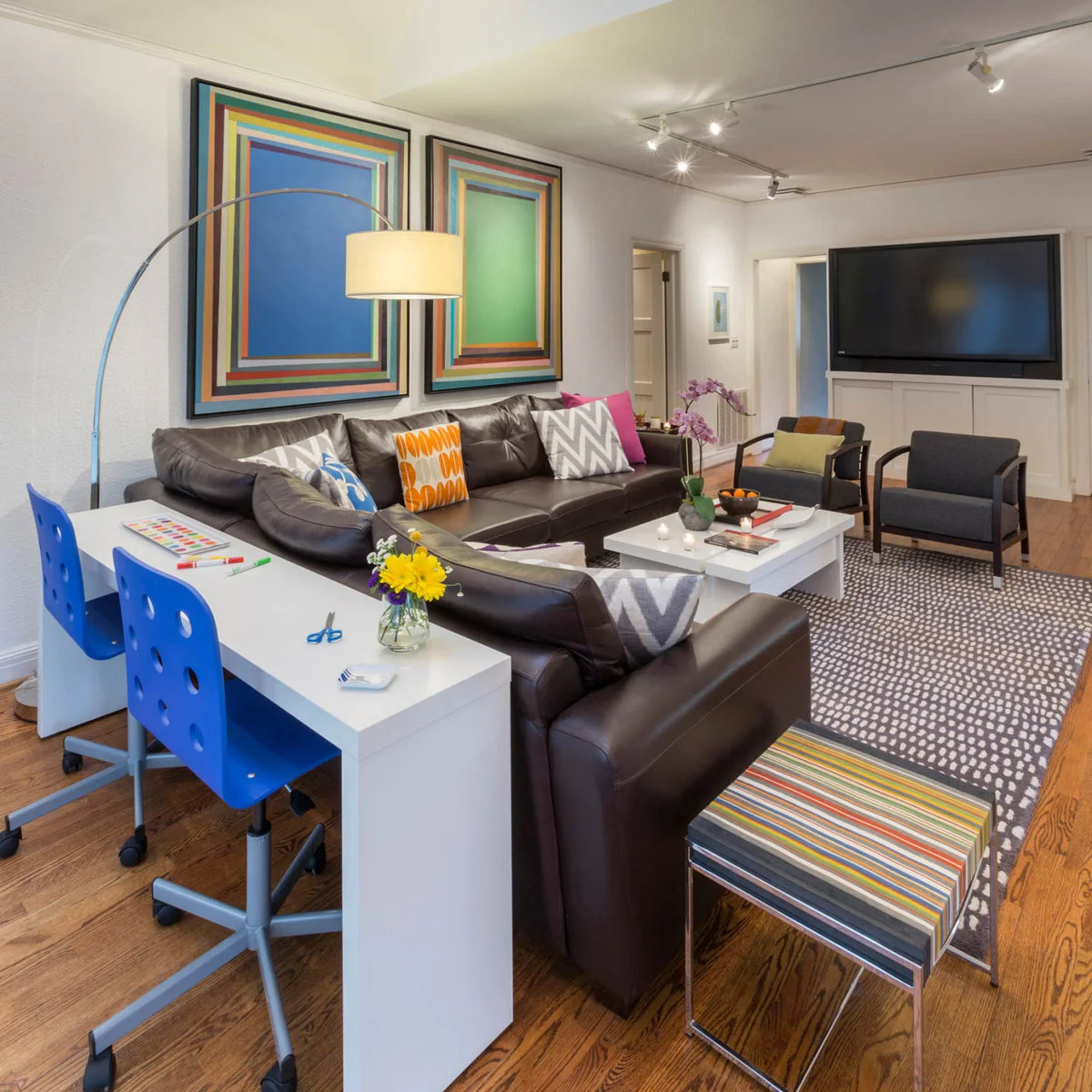 A long white desk with two blue chairs behind a dark sectional sofa in a colorful living room.