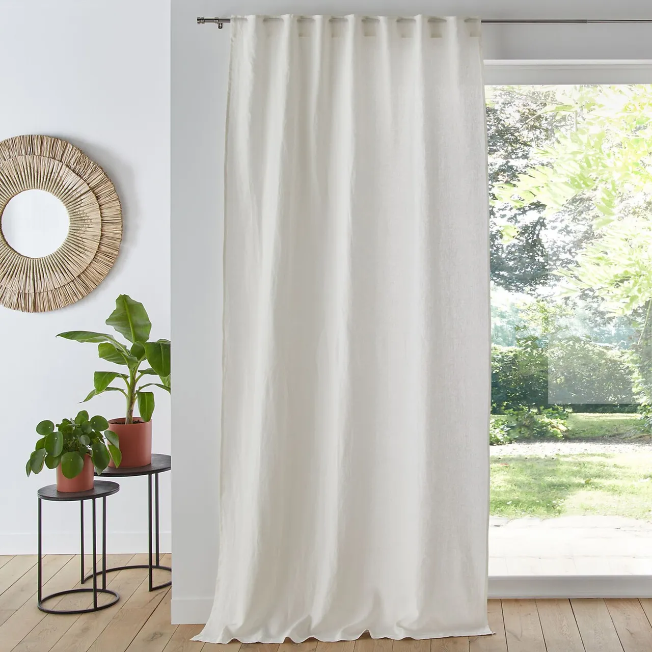 A pale room has long white curtains beside glazed doors, a round woven mirror and two small plants on slim black stands.