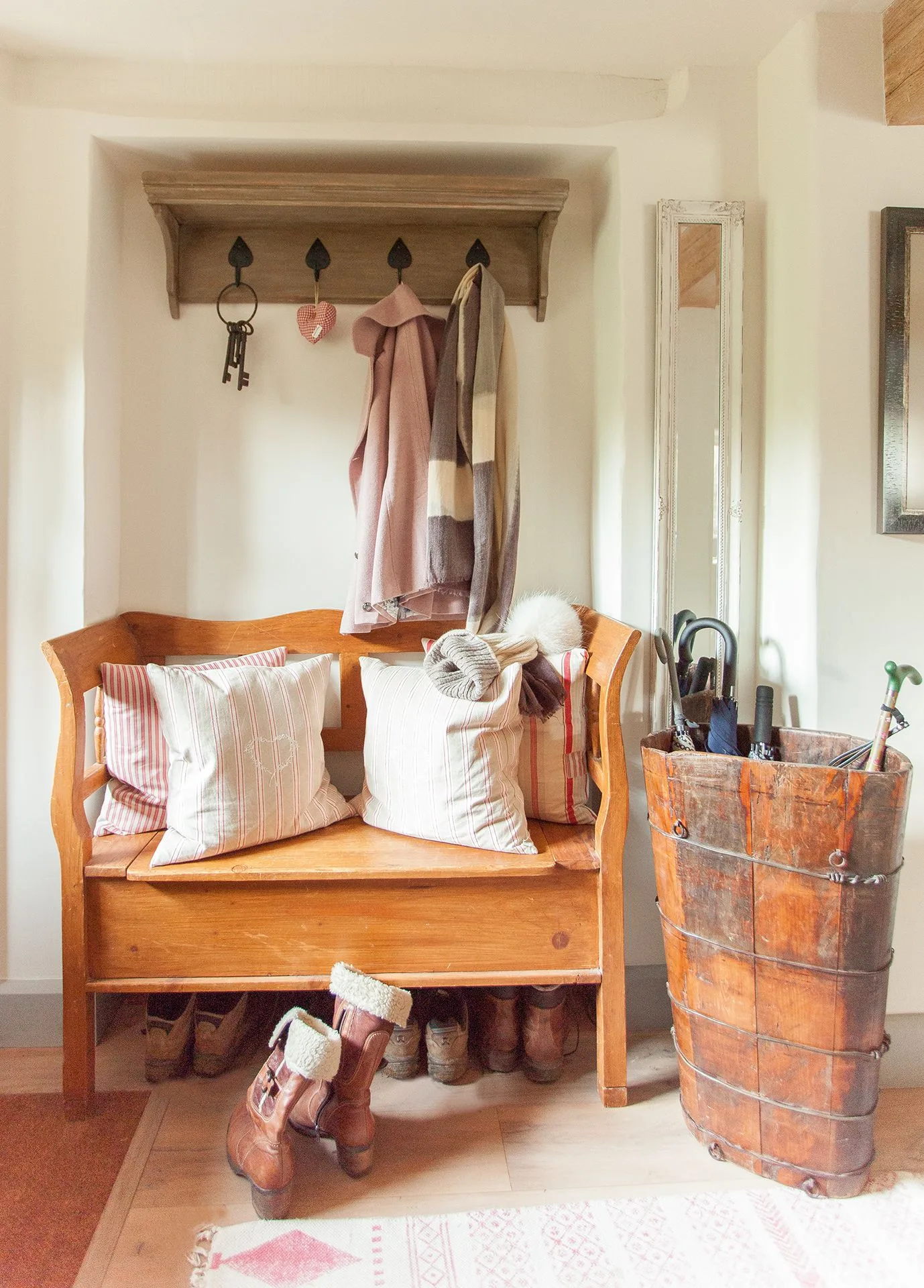 Entrance corner with a wooden bench, striped cushions, high wooden hook rail, shoes underneath and a tall wooden umbrella container.