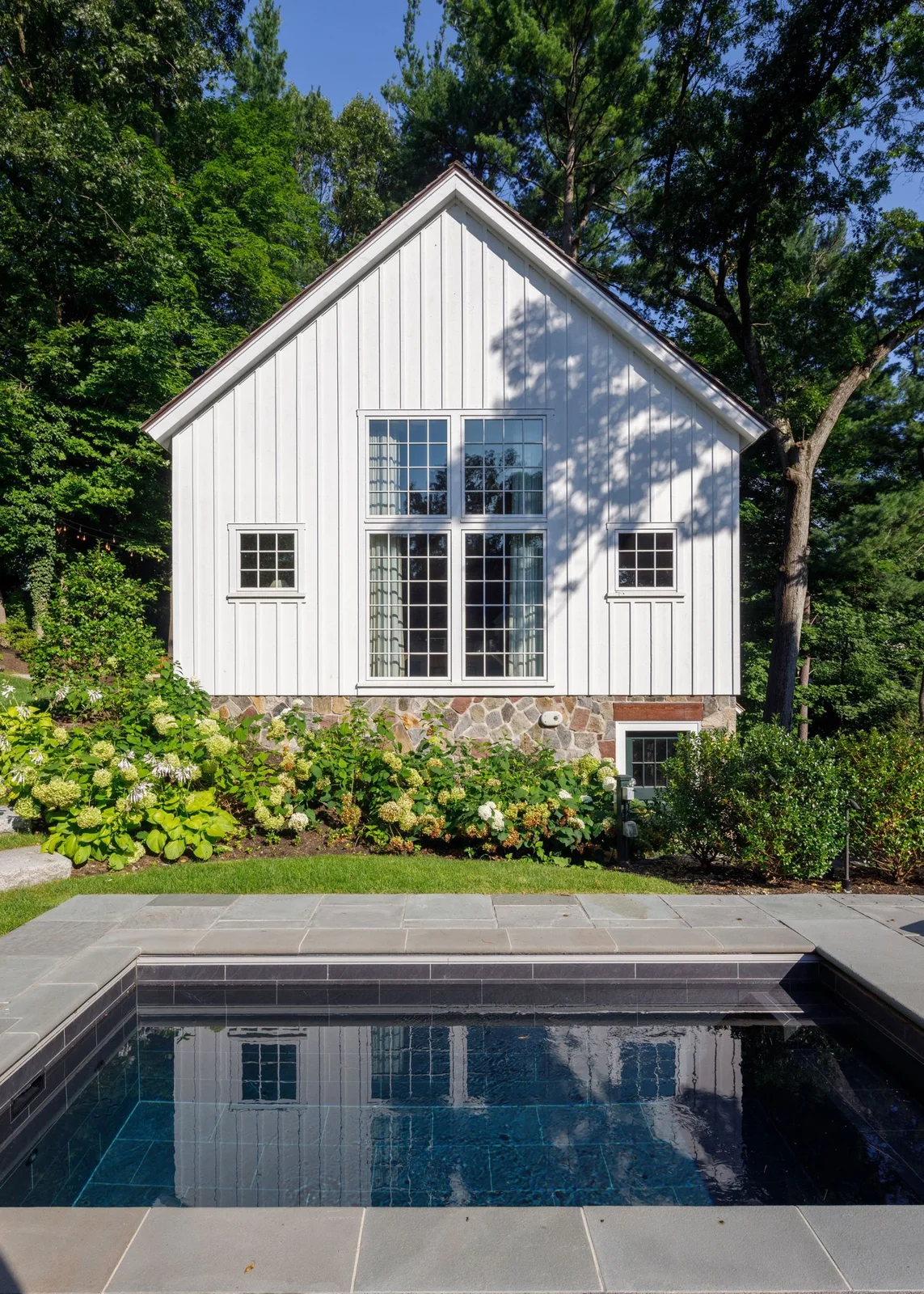 A small rectangular pool with a dark tiled interior and gray coping reflects a white gabled house and nearby trees.