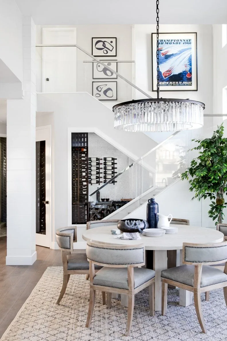 Recessed bottle storage beneath white stairs near a round white dining table, pale upholstered chairs, framed pictures, a plant, and a cylindrical hanging light.