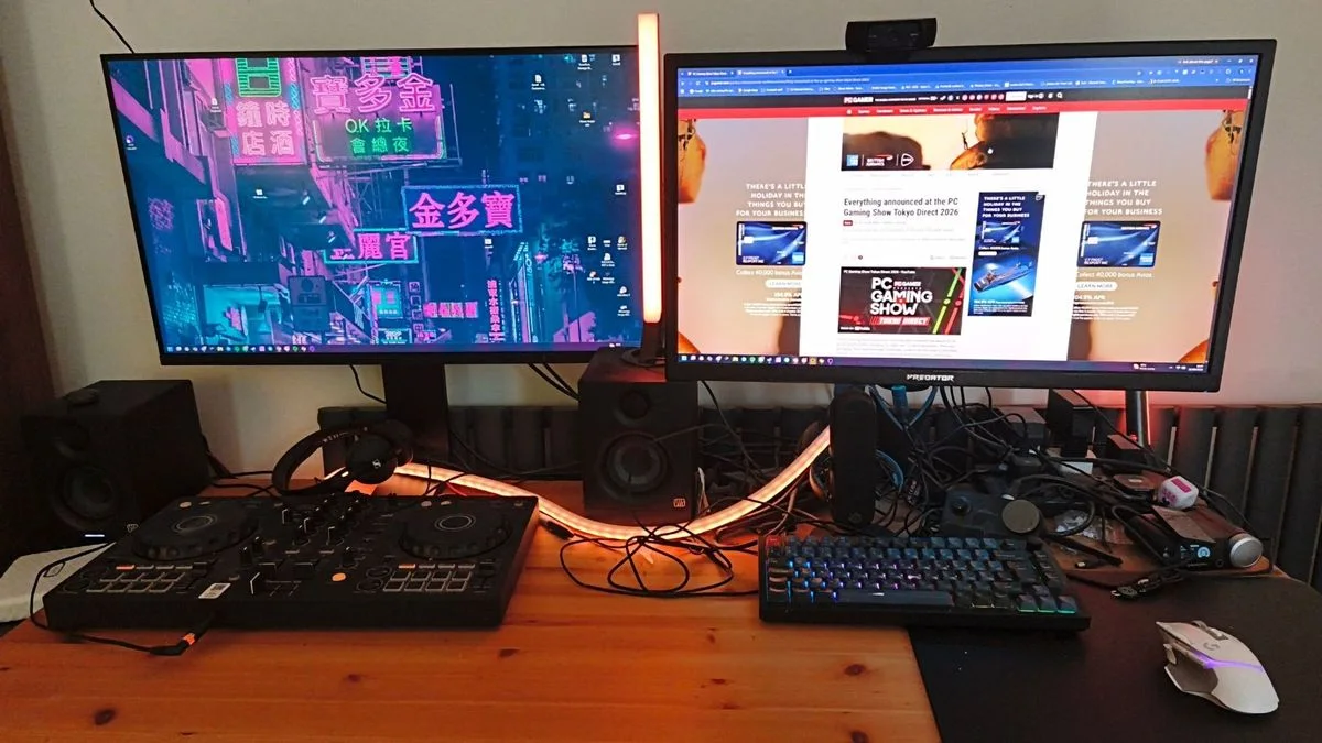 A wood desk with two monitors, a DJ controller, a keyboard, a microphone and visible cables between the equipment.