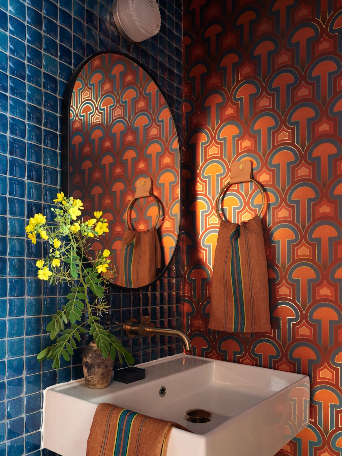 Orange geometric wallpaper meets glossy blue square tiles around a white wall basin, oval mirror, warm-metal towel ring and striped orange hand towels.