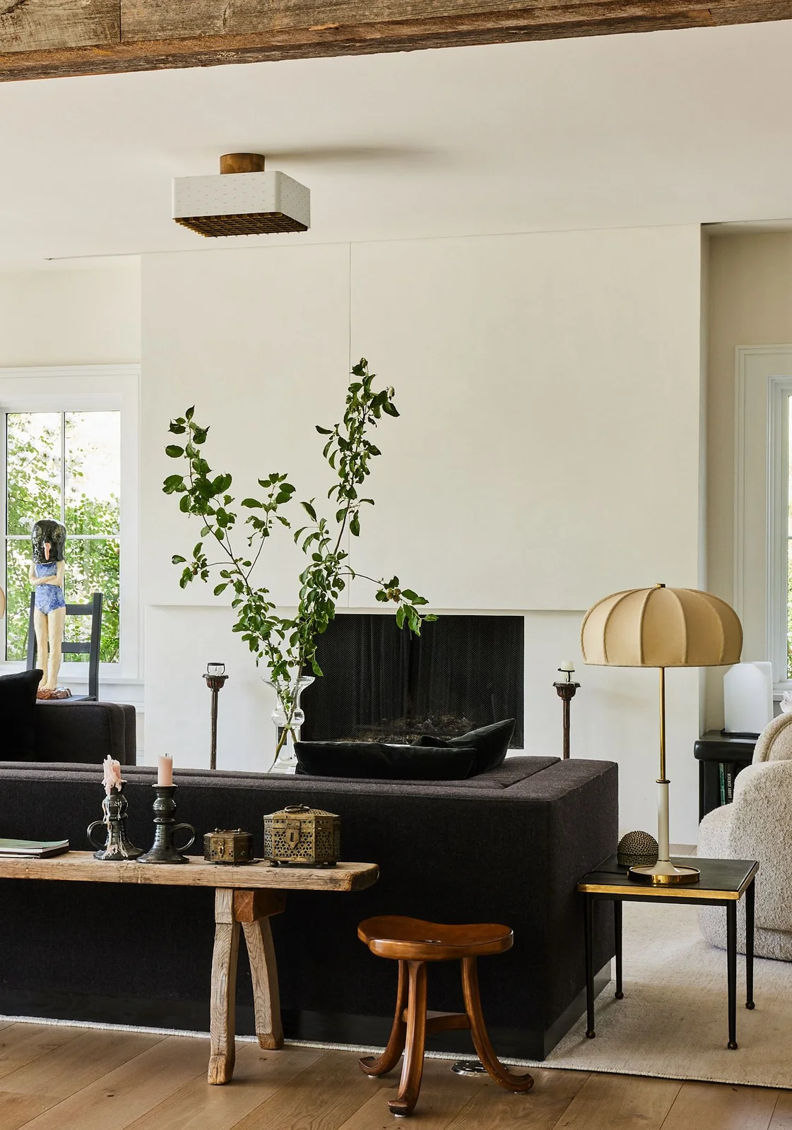 Pale panels conceal a television above a dark fireplace in a room with a dark sofa, wooden stool and gold-toned lamp