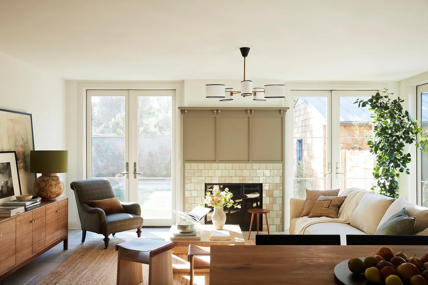 Pale cream square tiles frame a fireplace beneath muted painted paneling between glazed doors, with a pale sofa and wood furniture.