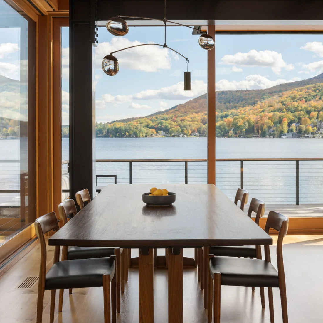 A dark rectangular dining table with slim wood chairs and black seats sits below a balanced globe light beside tall wood-framed windows and a water view
