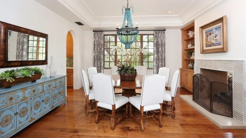 White dining room with a blue chandelier, blue decorative sideboard, white upholstered chairs and a fireplace.