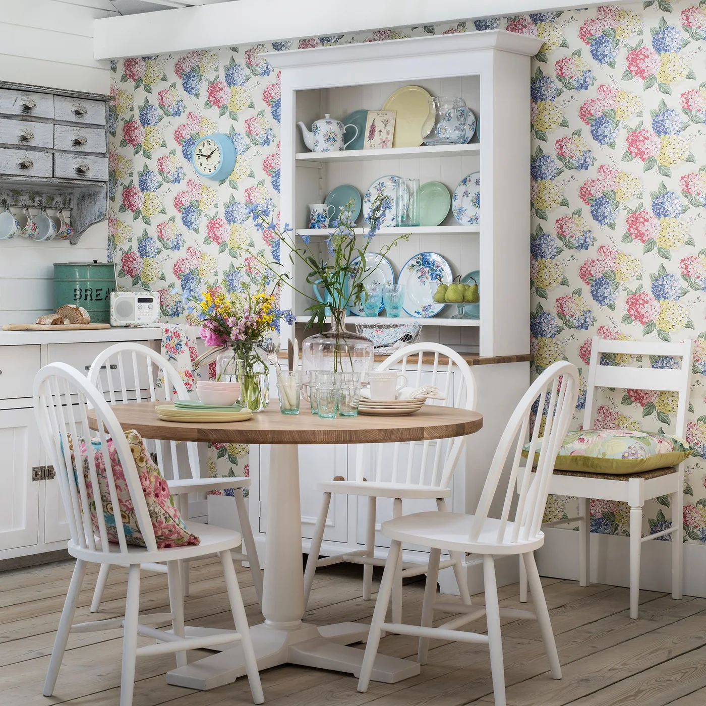 A white dresser displays floral plates and pitchers above lower cupboards beside a round dining table and flower-patterned wallpaper.