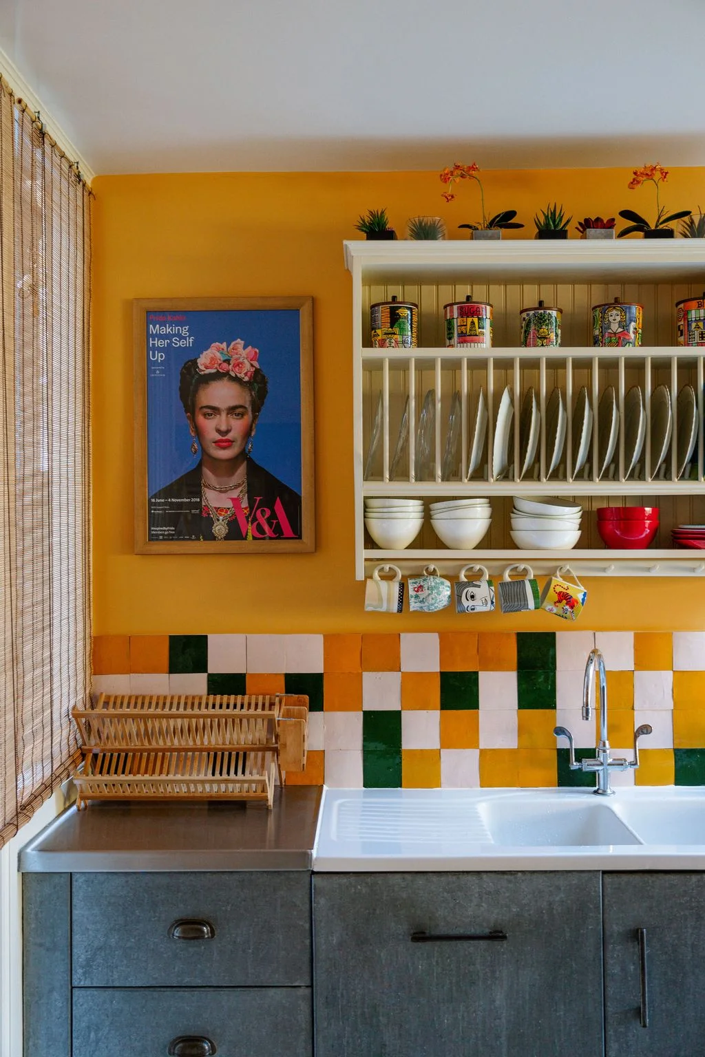 A white wall rack holds plates, mugs and colorful tins above a sink with a yellow wall, mixed square tiles and framed artwork.