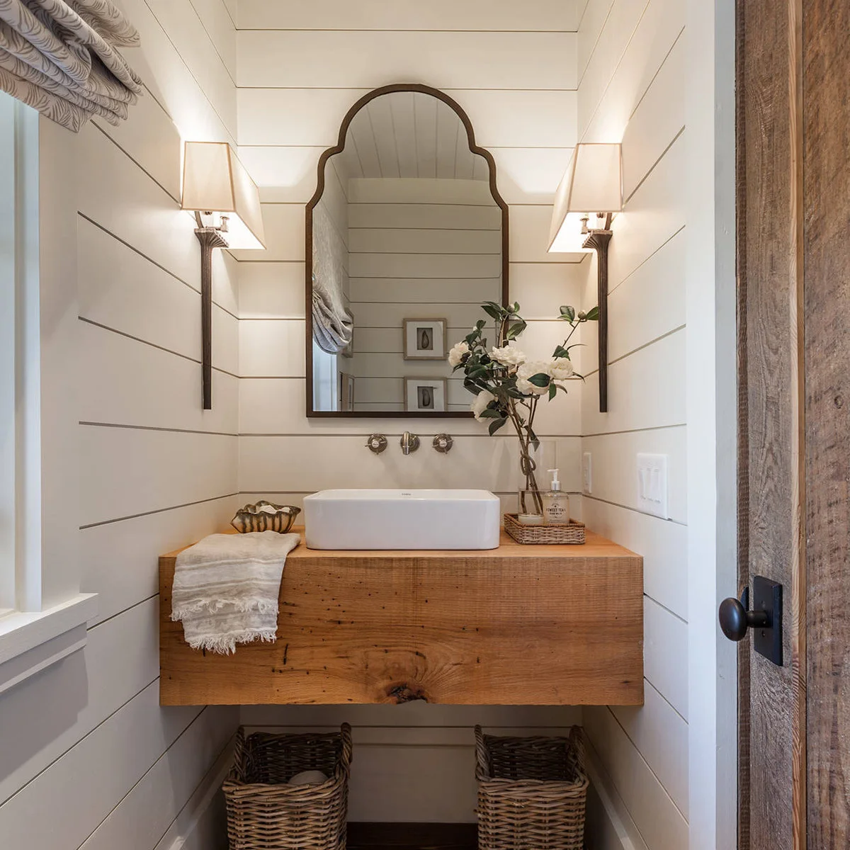 Tall dark-stem sconces with pale tapered shades sit on side walls beside a shaped mirror over a vessel sink.