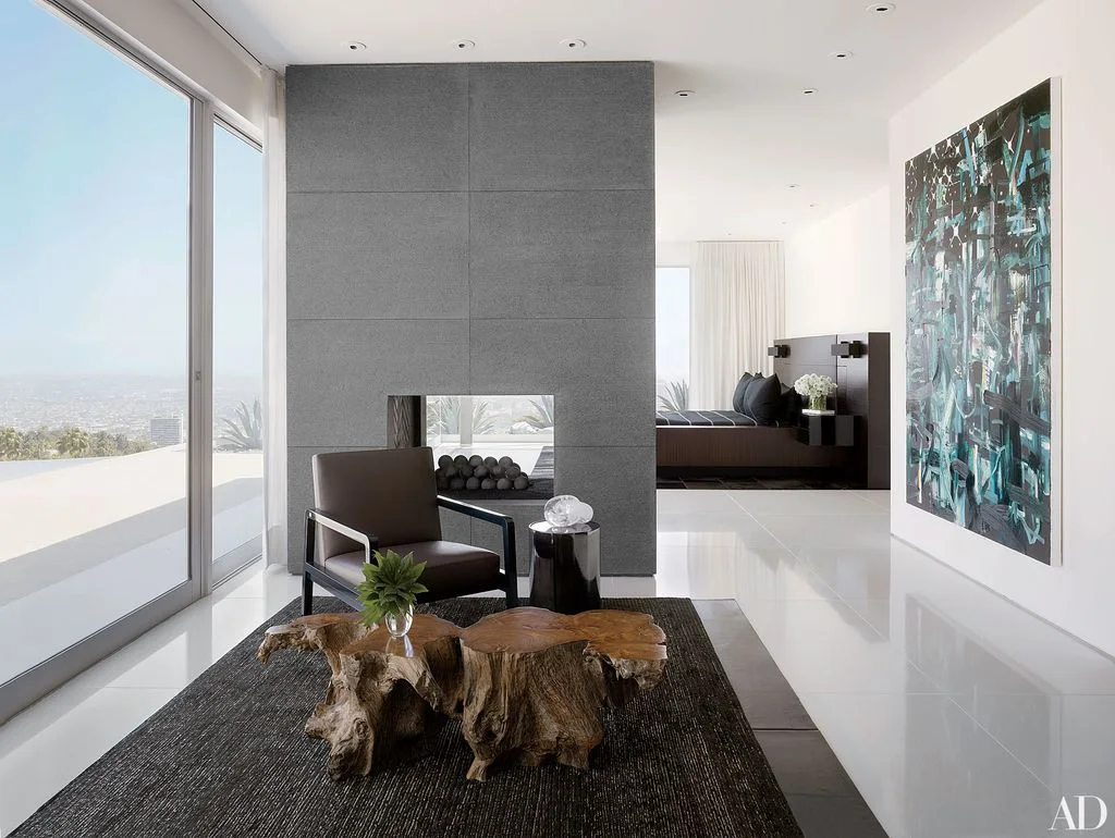 A gray paneled fireplace partition has a small rectangular opening beside a glazed wall, dark chair, textured wood coffee table and a separate sleeping area