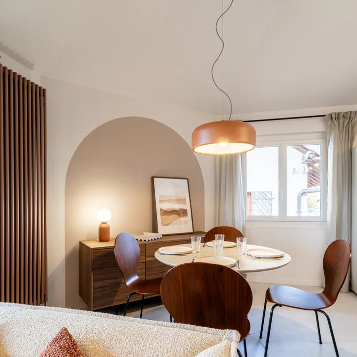 A round white table and curved wood chairs stand before a low wood sideboard and a muted arched wall shape.