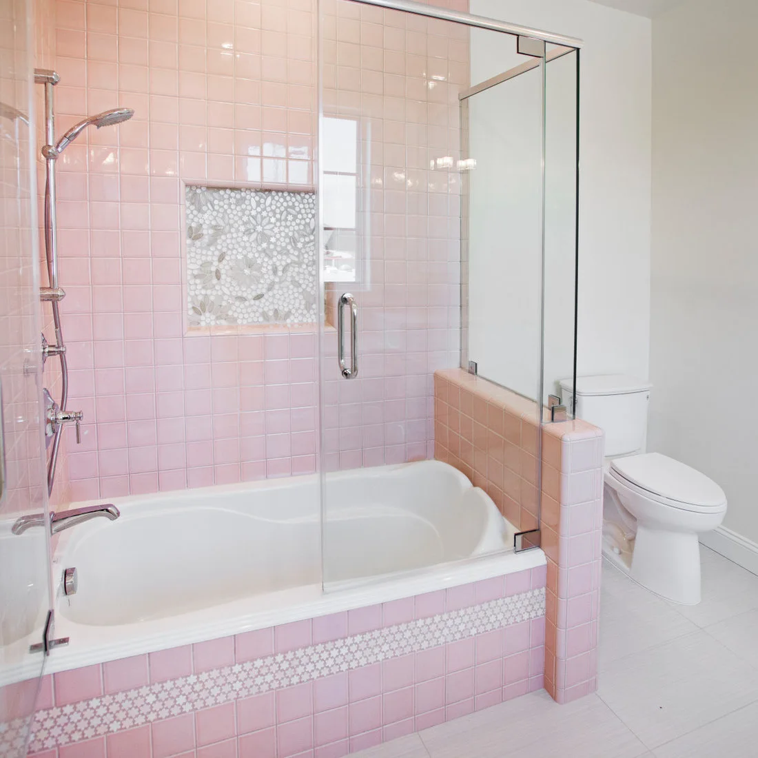 Pink square tiles surround a white bath with a mosaic band across the bath panel and inside a recessed niche