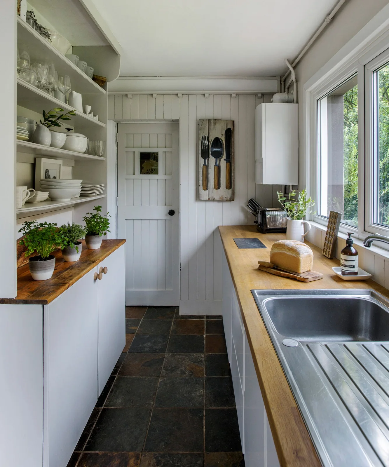 Compact white galley kitchen with wooden counters and open shelves opposite a window sink