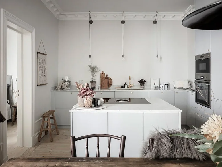 A pale kitchen has flat cabinet fronts, a small island with a cooktop, simple suspended bulbs and a wood dining table in the foreground.