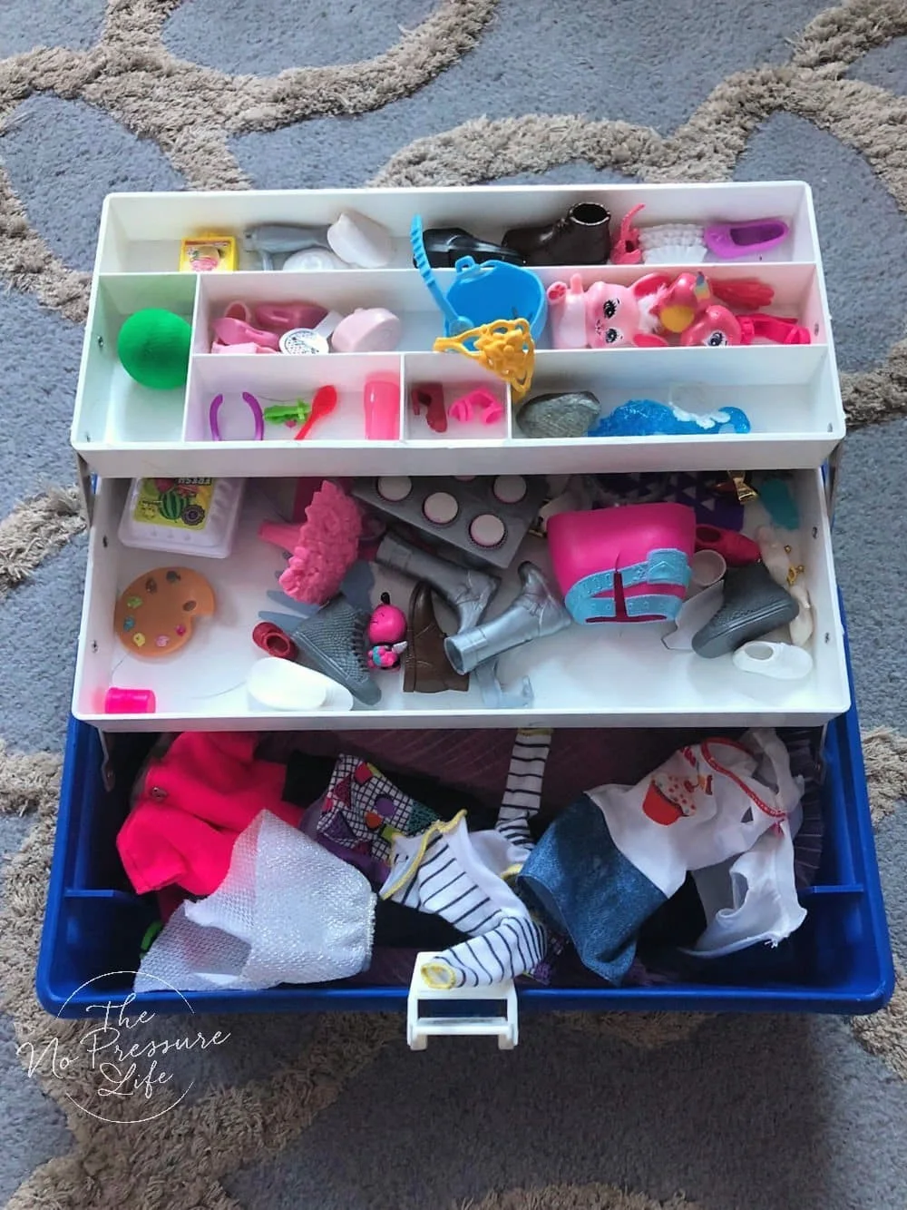 Blue storage box with two white cantilever trays containing small doll accessories above clothing in the base.