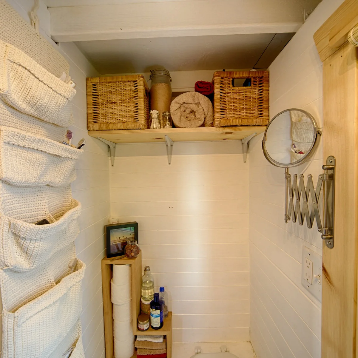 Tiny wood-lined bathroom with a shelf of baskets, hanging fabric pockets and a slim toilet-paper cubby