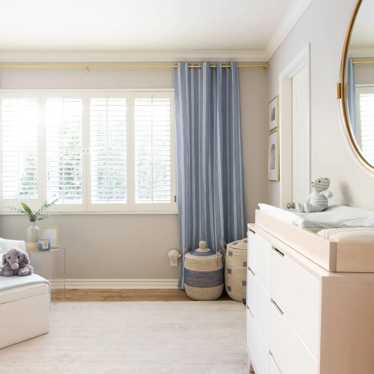 White nursery dresser with a changing tray beside blue striped curtains and woven baskets