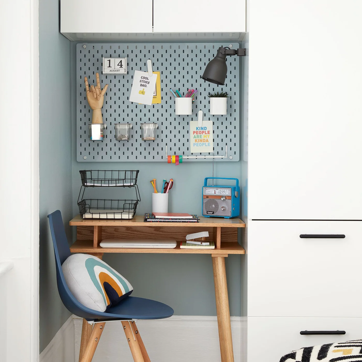 A small wood desk fits beside white wardrobe drawers beneath a blue-gray pegboard with containers, paper trays, a black light and a blue chair.