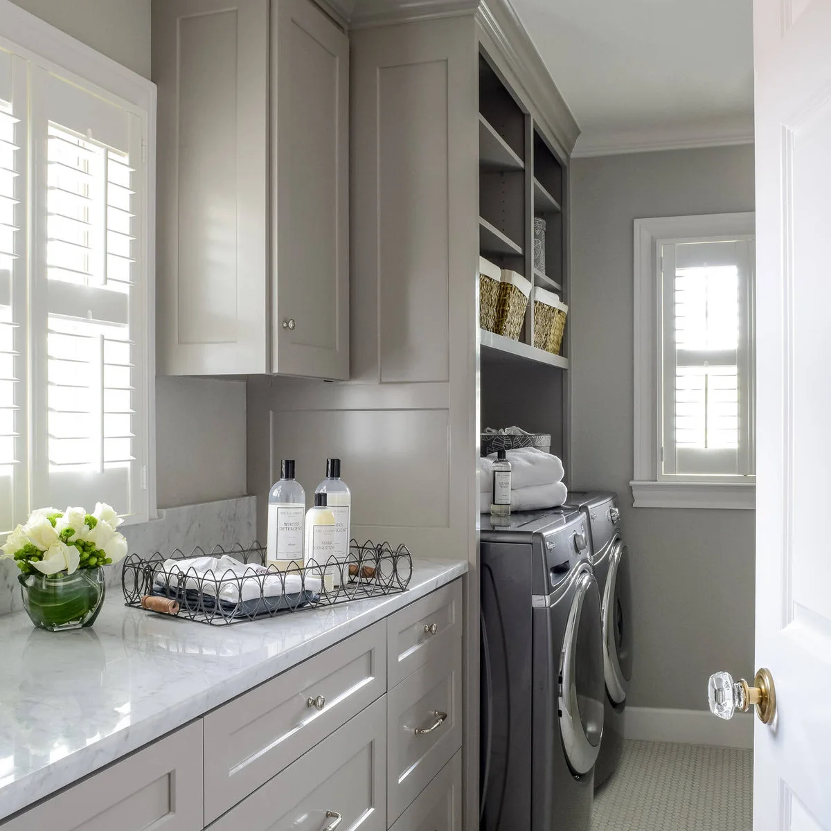 Narrow pale laundry room with a long left counter, machines at the far end, shuttered windows and open shelves with baskets