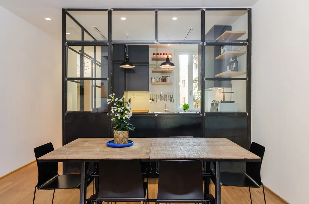 Black-framed glass separates a dark kitchen from a dining table with black chairs.