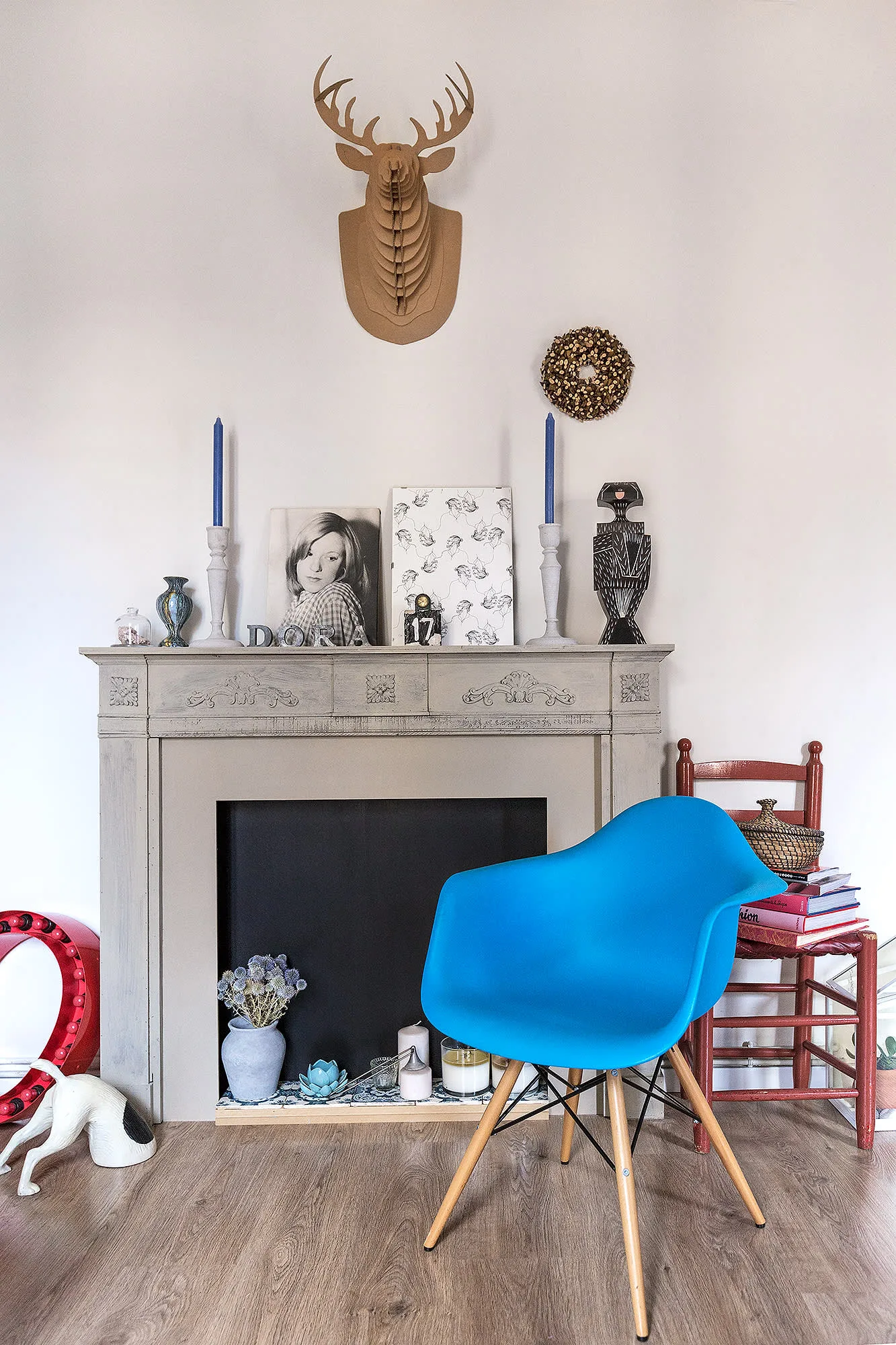 Pale decorative fireplace with blue tapers in pale holders, portraits above and a bright blue chair in front.