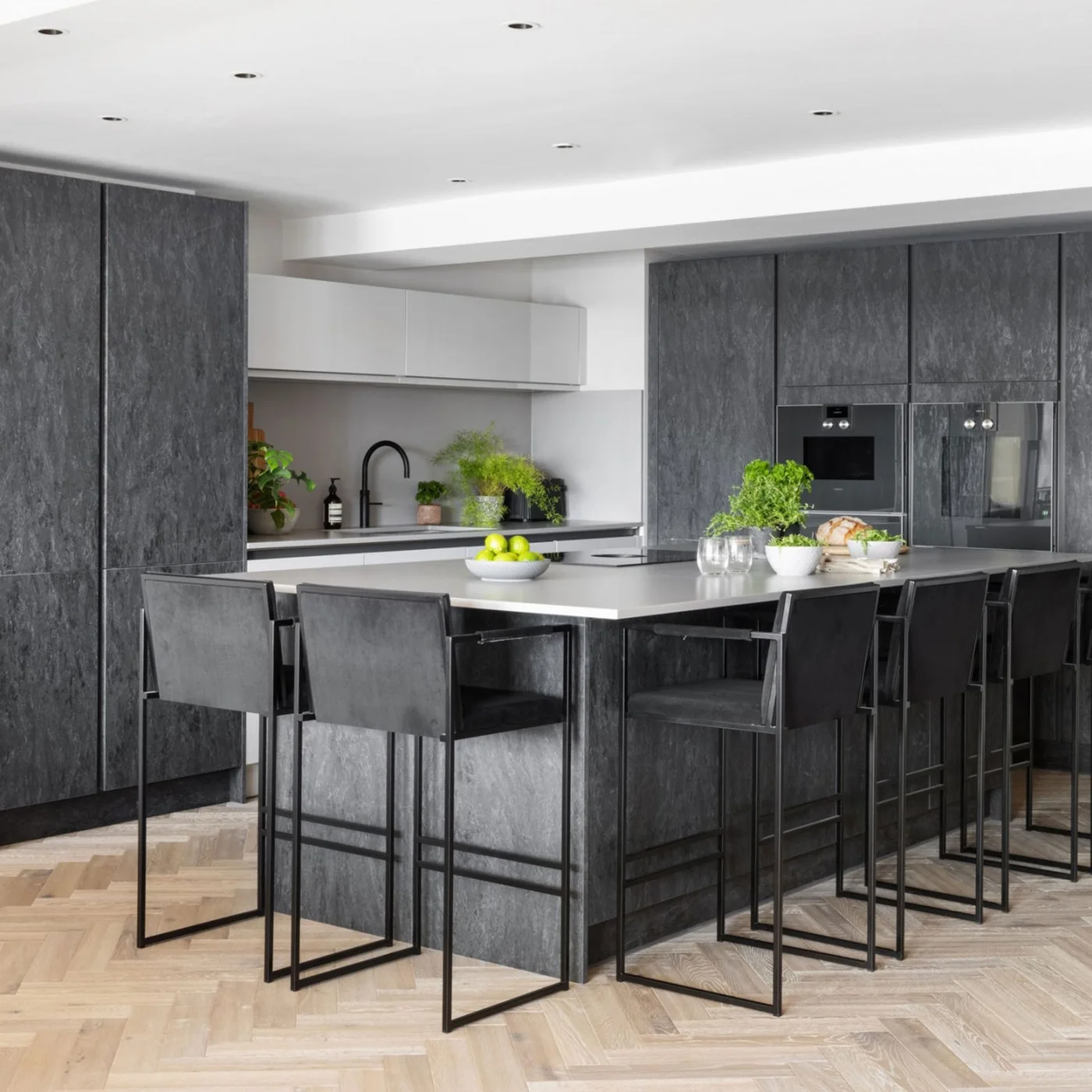 Flat dark textured cupboards surround built-in kitchen appliances behind a matching island with four open-framed dark stools and a pale counter