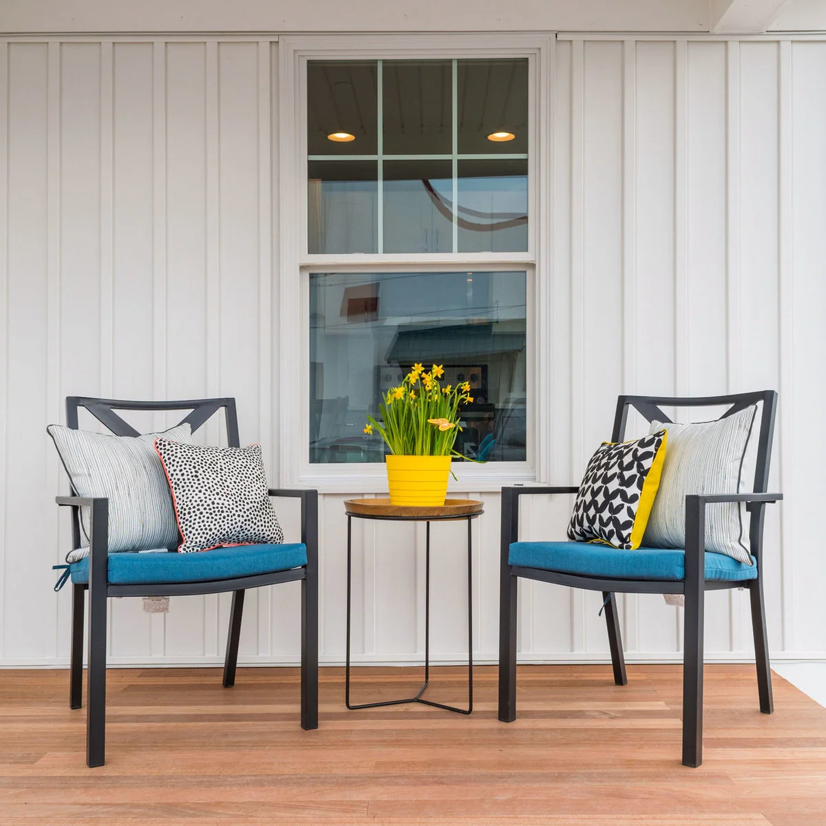 Two dark upright porch chairs with blue cushions and a small round table beneath a window