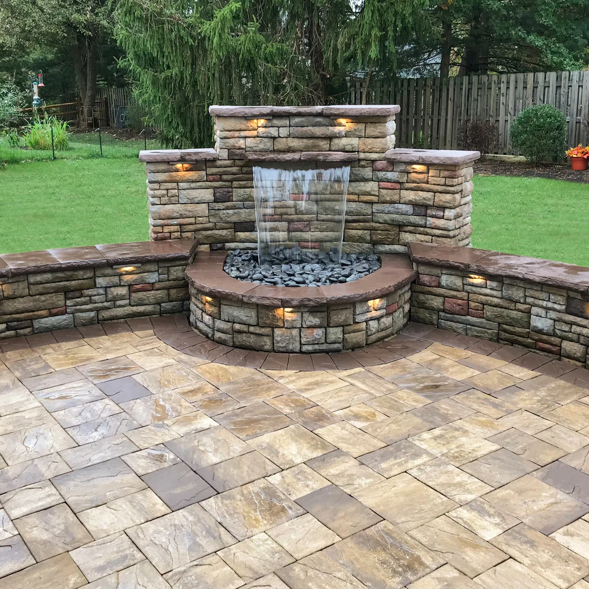 A compact block-faced wall with one broad sheet of falling water, a semicircular stone-covered basin and small integrated lights.