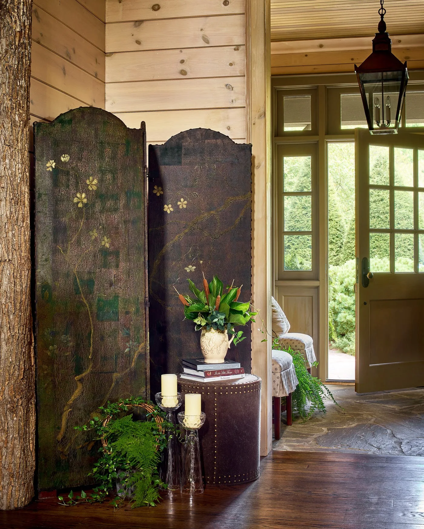 Dark candle lantern near wood glazed door in timber-lined entry with decorative folding screen, textured floor and grouped flowers
