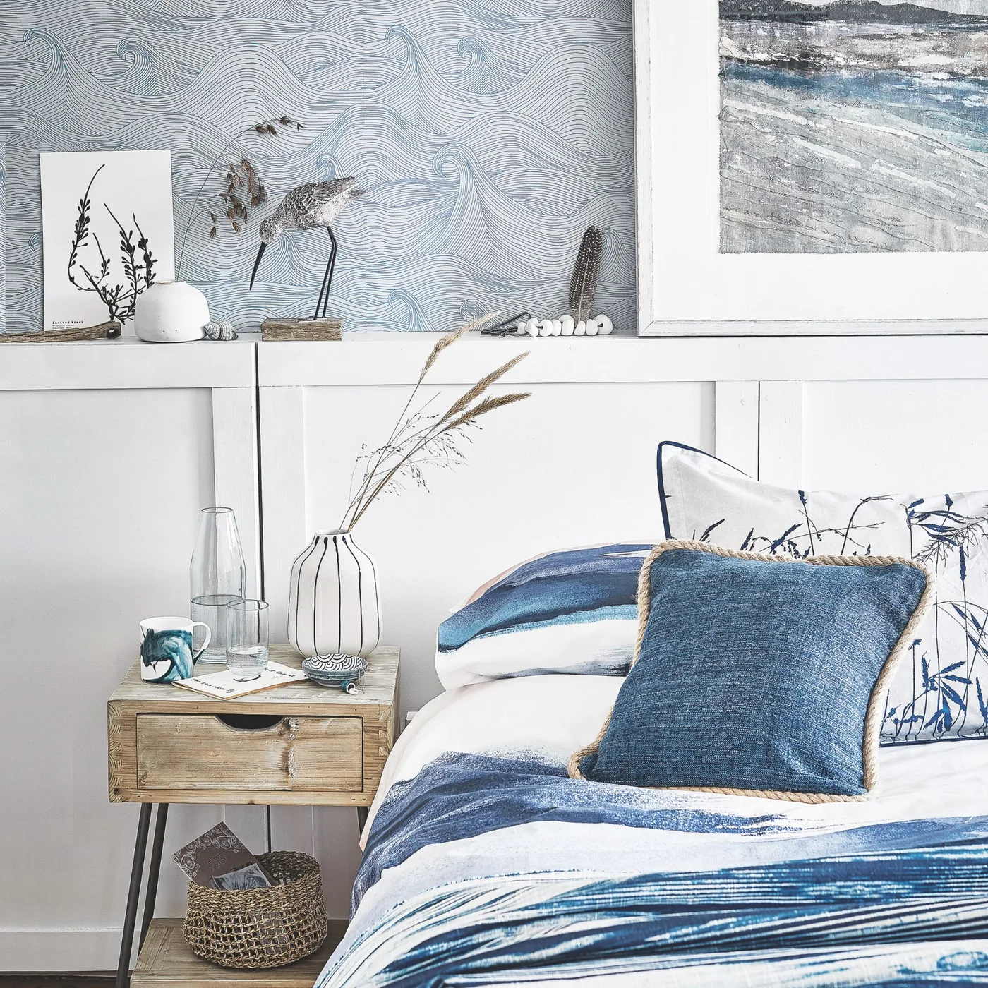 Bedroom detail with pale wave-pattern wallpaper, white lower paneling, blue-and-white bedding and a small wooden bedside drawer.