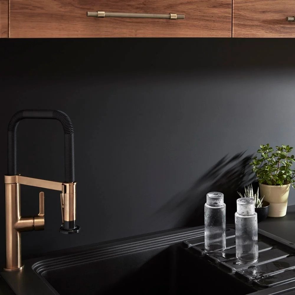 A black kitchen sink and dark backsplash sit below wood-fronted storage beside a black-and-gold-colored flexible tap and small glass bottles