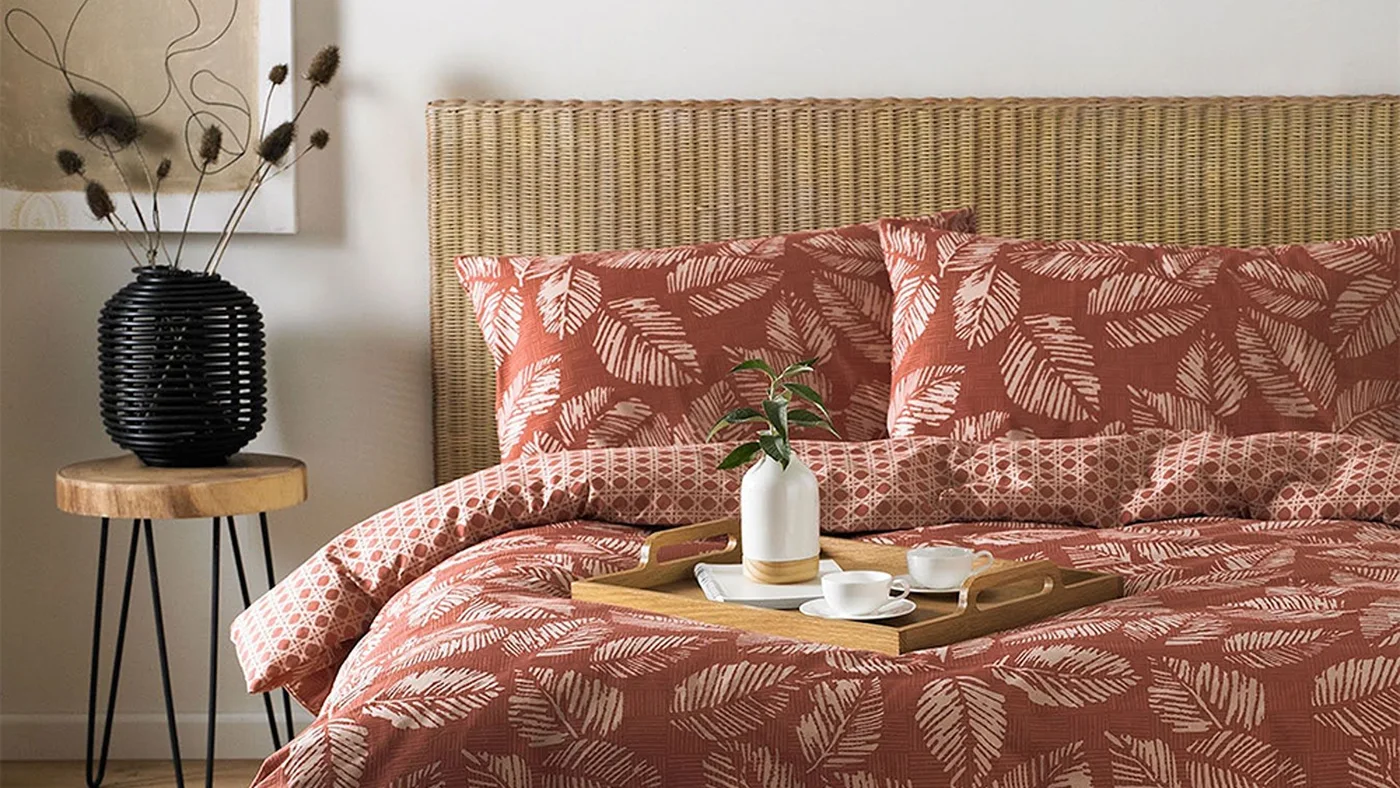 Red-brown bedding with a pale leafy print sits before a woven headboard beside a black ribbed vase on a small wooden table