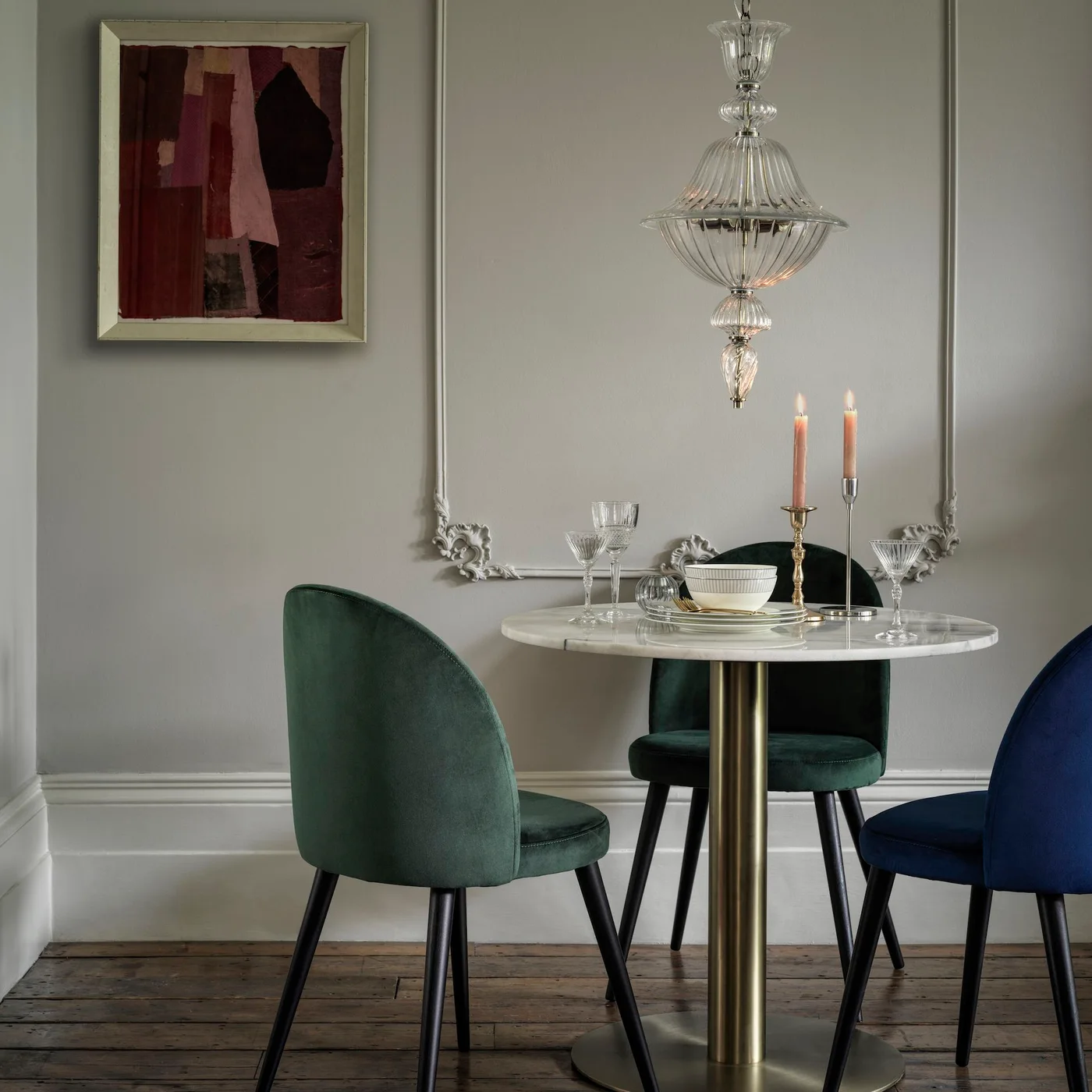 Green and blue upholstered chairs surround a pale round pedestal table beneath a glass pendant against gray walls