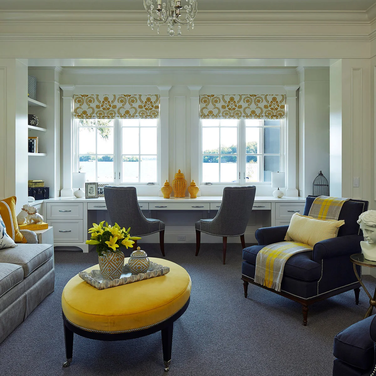 Two chairs at a long fitted desk beneath windows, behind a living room seating group with yellow accents.