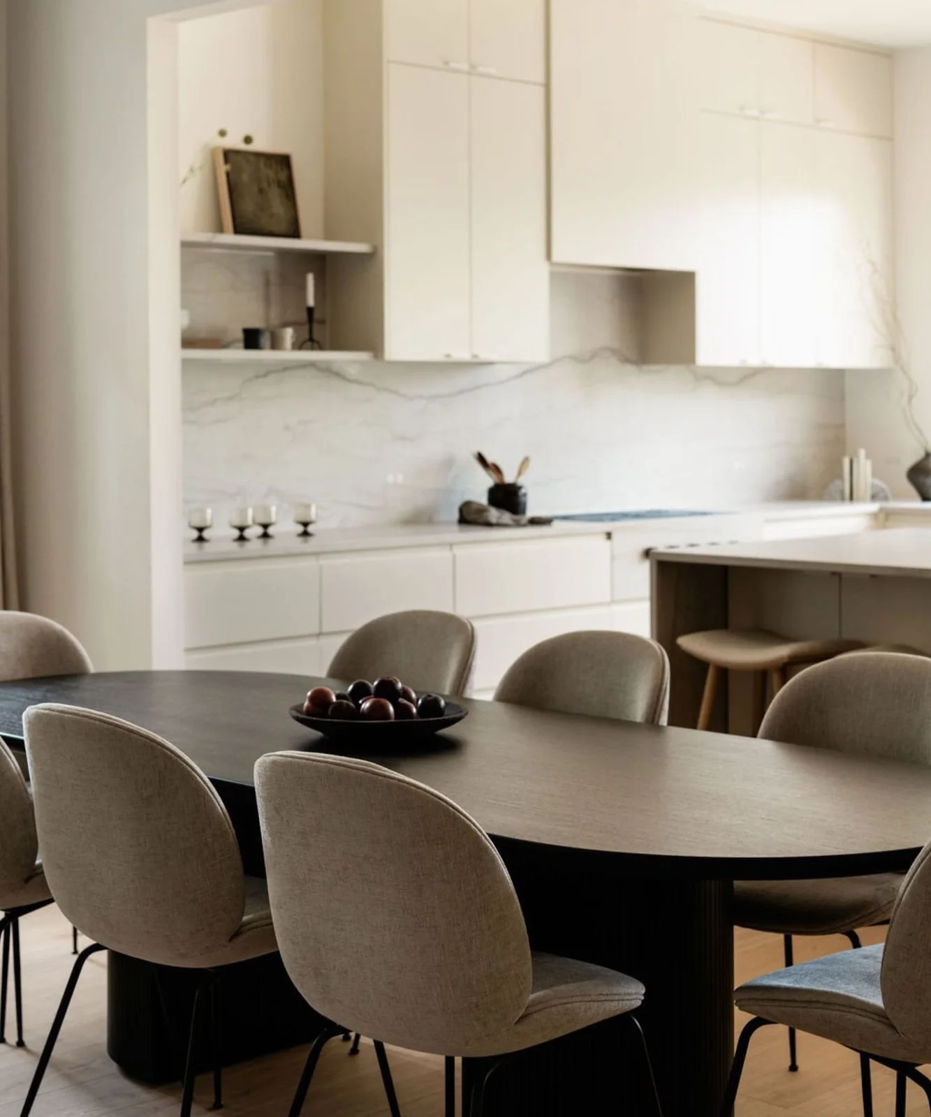 A dark rounded dining table and soft gray chairs stand beside pale kitchen cabinets, a veined backsplash and wooden stools