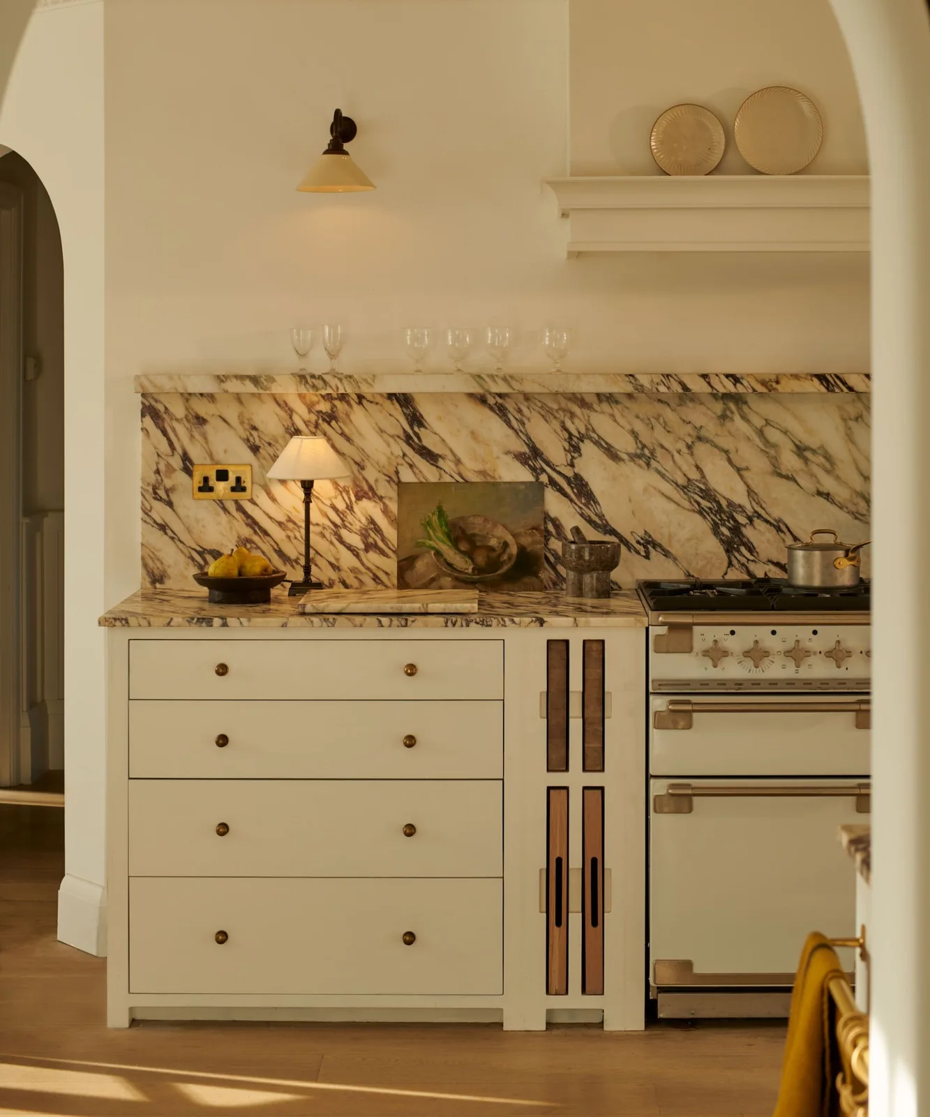 Warm pale drawers and small dark knobs sit beneath a strongly veined backsplash beside a cream range.
