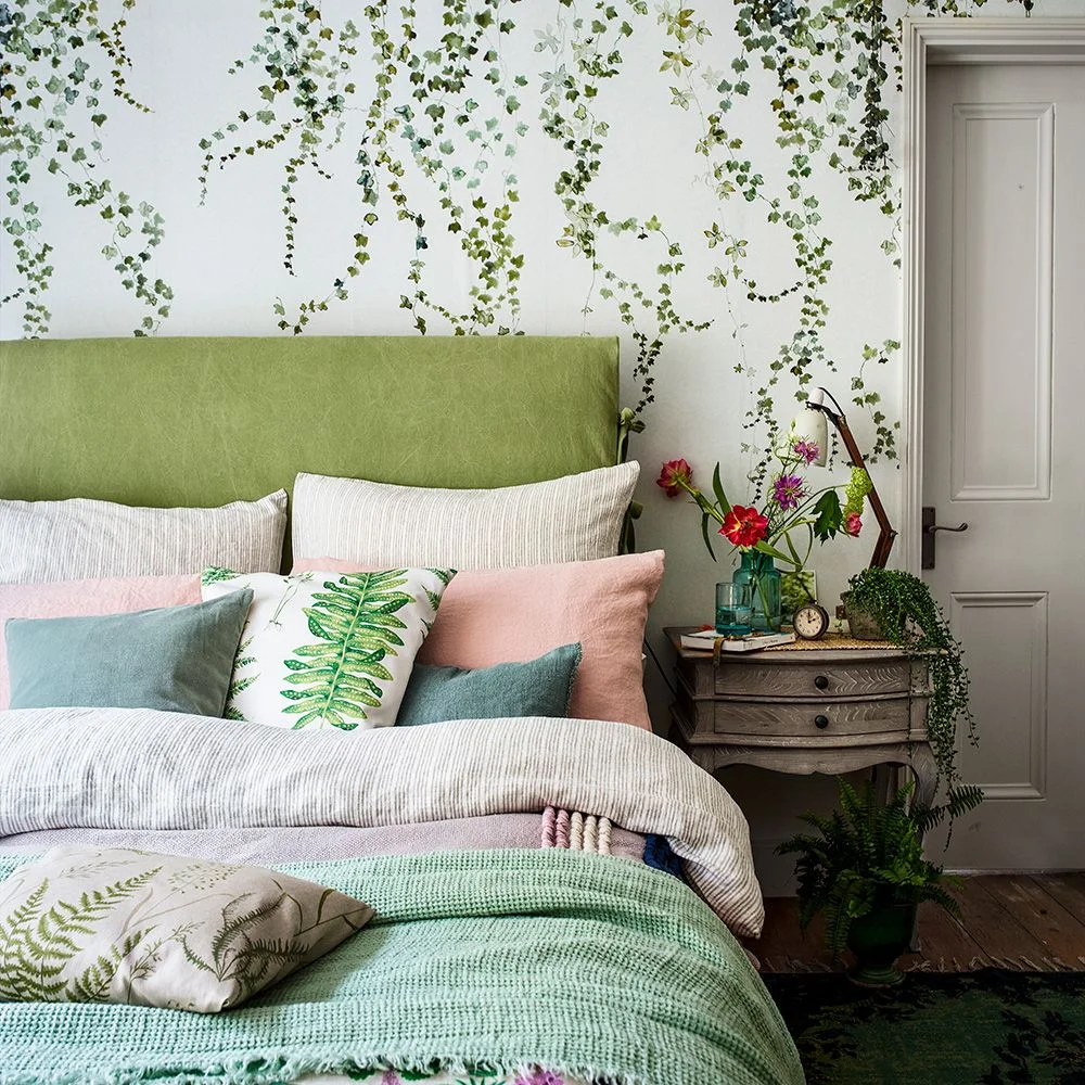 Green trailing-leaf wallpaper surrounds an olive headboard with pale bedding, pink and blue-green cushions and a small bedside drawer unit