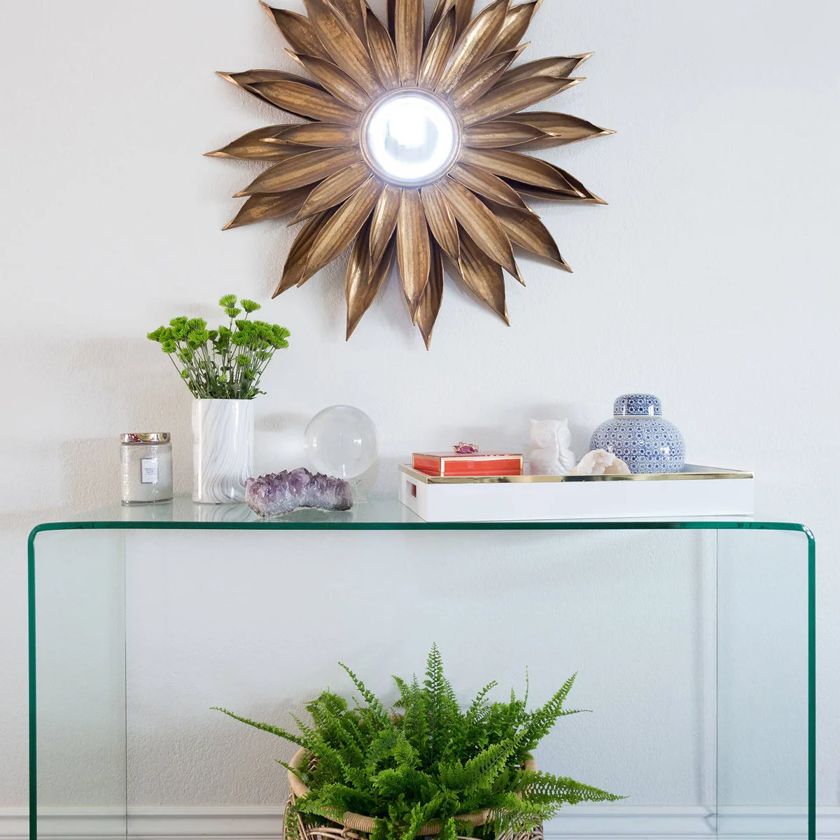 A clear console beneath a sunburst mirror with a tray of small objects, flowers and a fern in a basket below.