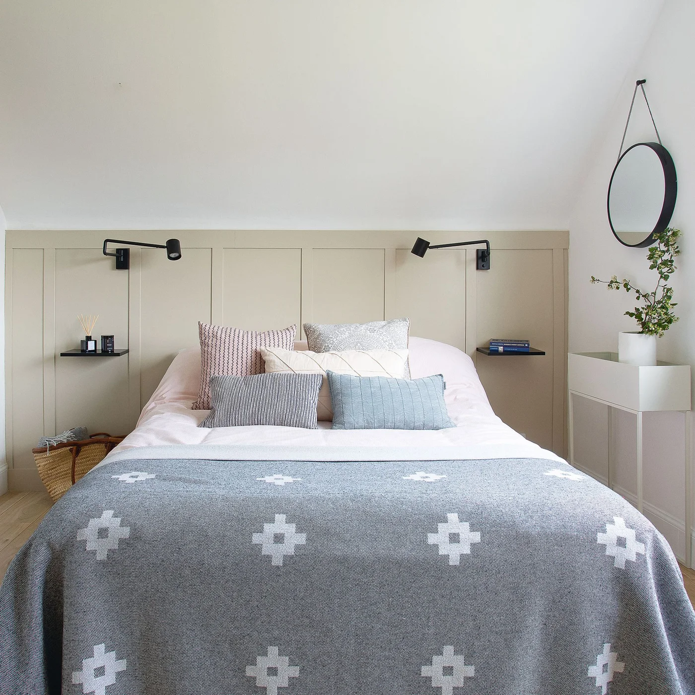 Two small black shelves and adjustable black wall lights flank a bed against pale lower-wall paneling, with a round mirror and slim white drawers nearby.