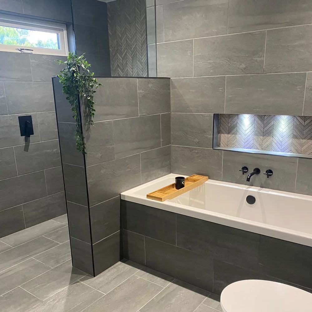 Gray tiled bathroom with a charcoal bath front, half-height shower partition, white rectangular tub, black taps, and illuminated niche.