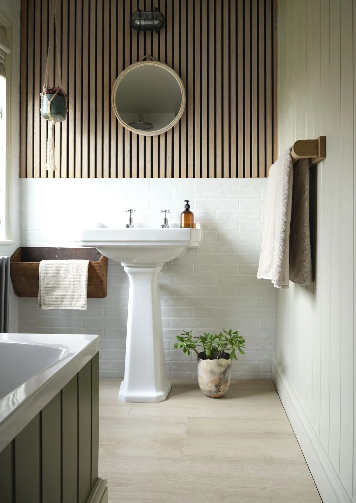 A white pedestal basin stands below wood slats and a small round mirror beside pale rectangular tiles, muted green paneling and a wood towel holder