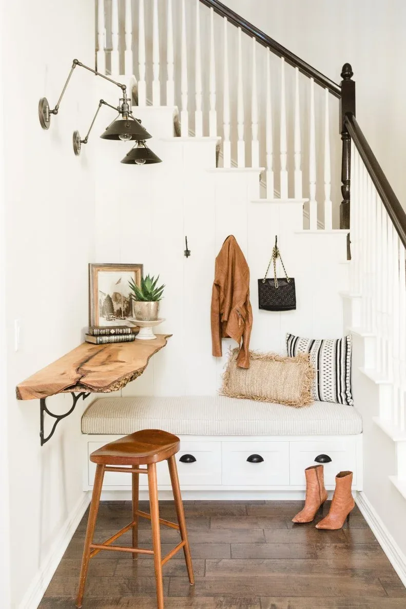 Under-stair entry nook with pale drawer bench, wooden side shelf, wall lights and striped cushions.