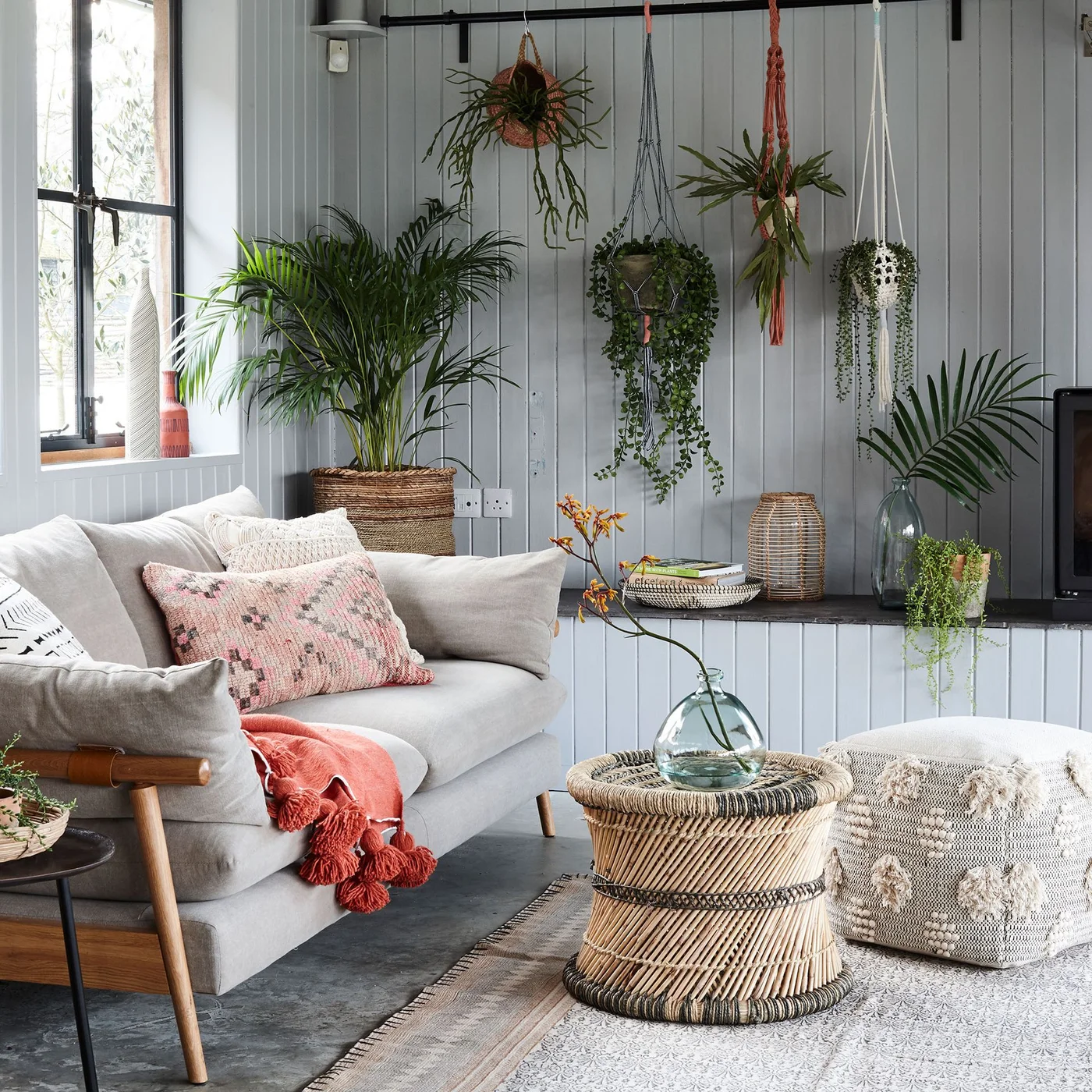 Gray vertical wall panels sit behind a pale sofa, hanging plants and a woven round coffee table