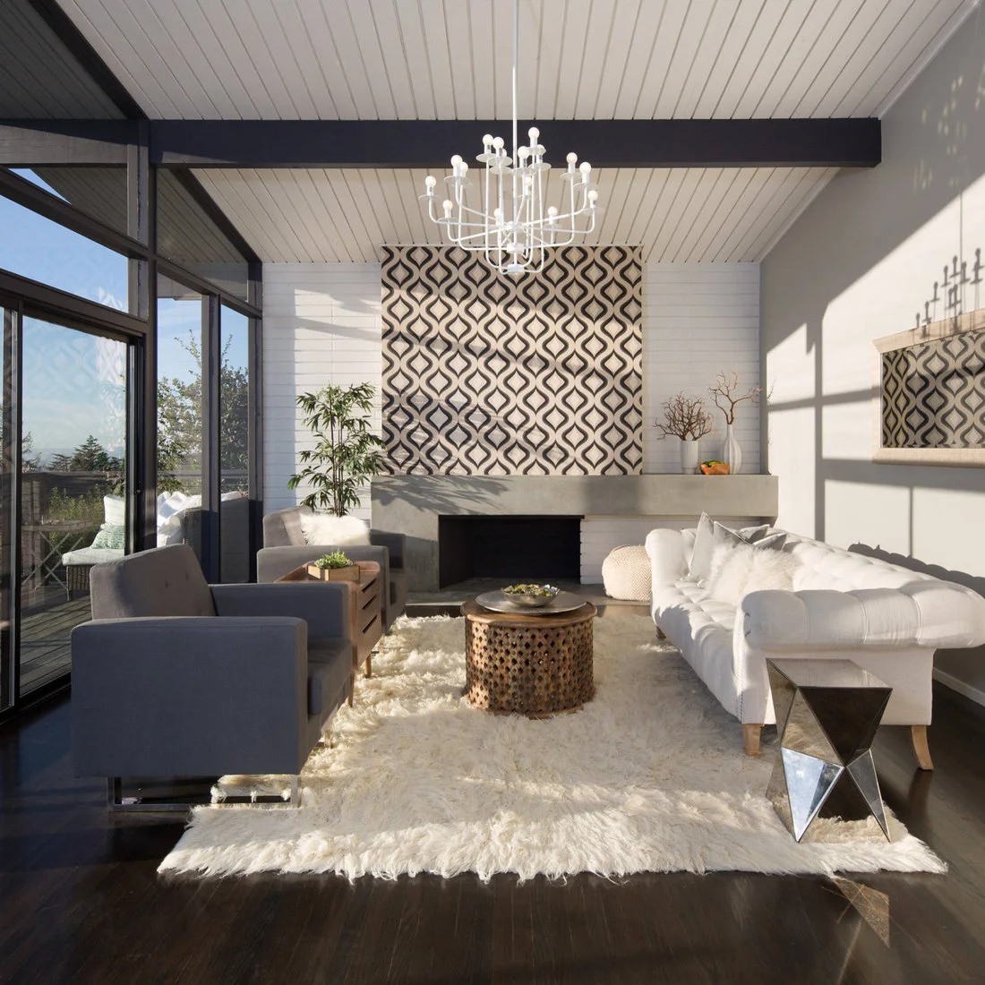 A pale living room has a broad simple fireplace beneath a black-and-cream geometric patterned chimney wall, with gray chairs, a white sofa and a pale textured rug.