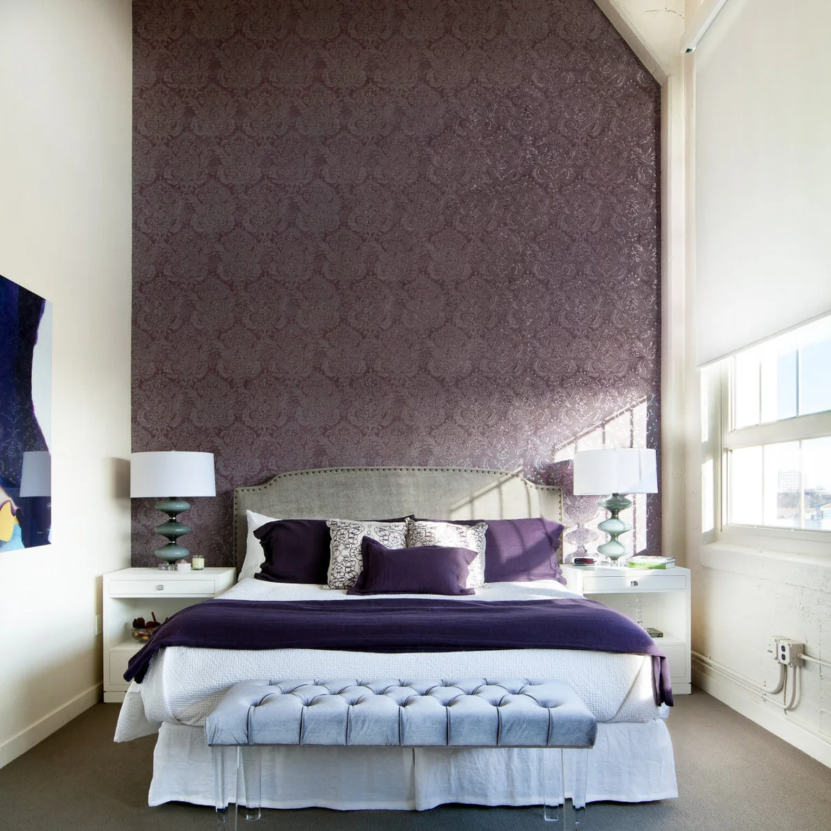 Purple patterned wallpaper behind a pale curved headboard on a tall bedroom wall with purple bedding.