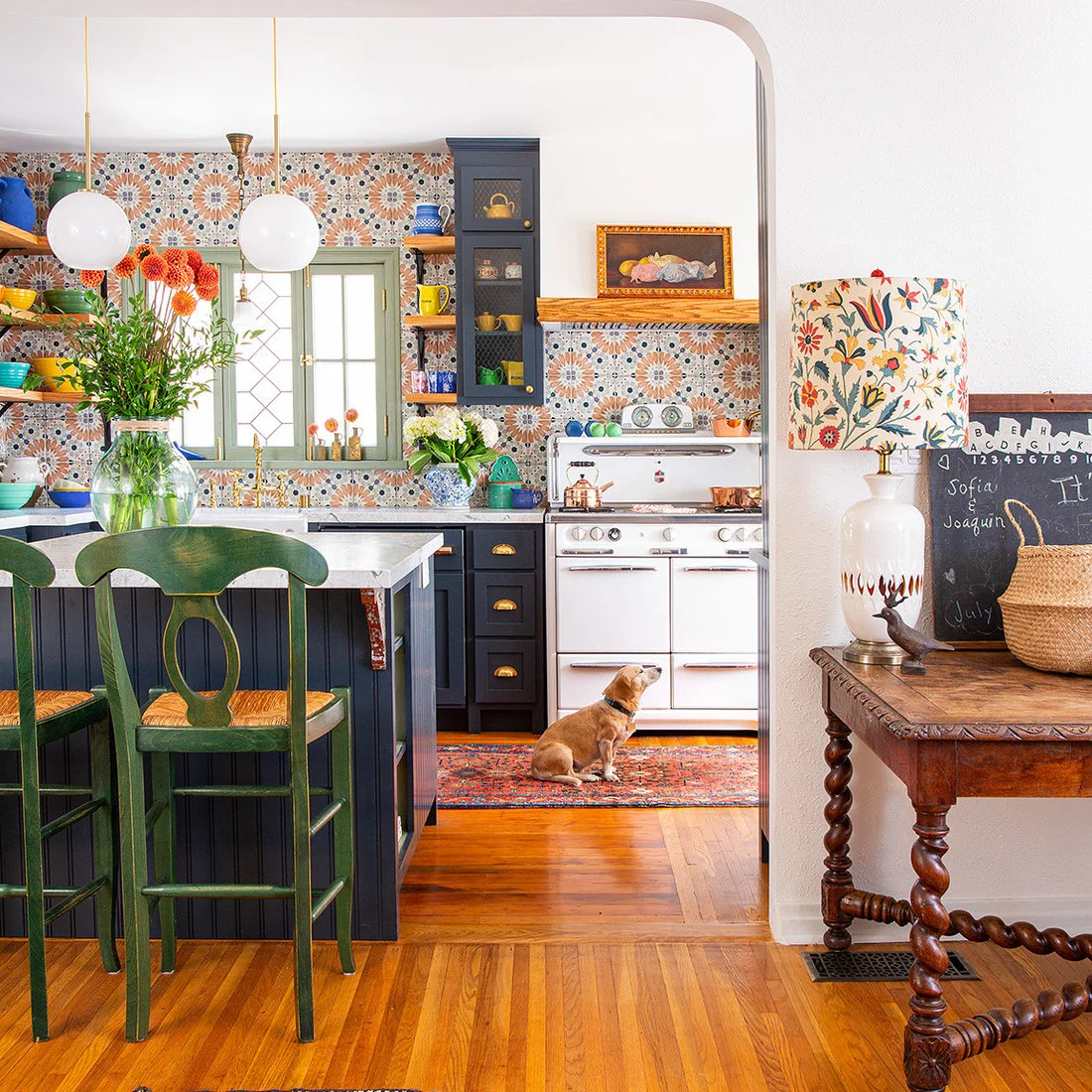 Colorful patterned tiles sit behind dark cabinets, green stools and open shelves in a kitchen framed by an arch.