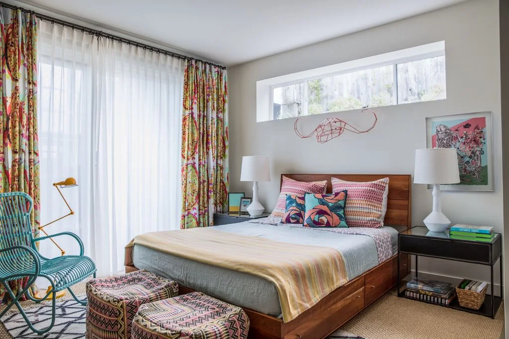 A simple wood bed stands beside dark open bedside units, a turquoise woven chair and colorful curtains with patterned cushions and small upholstered stools